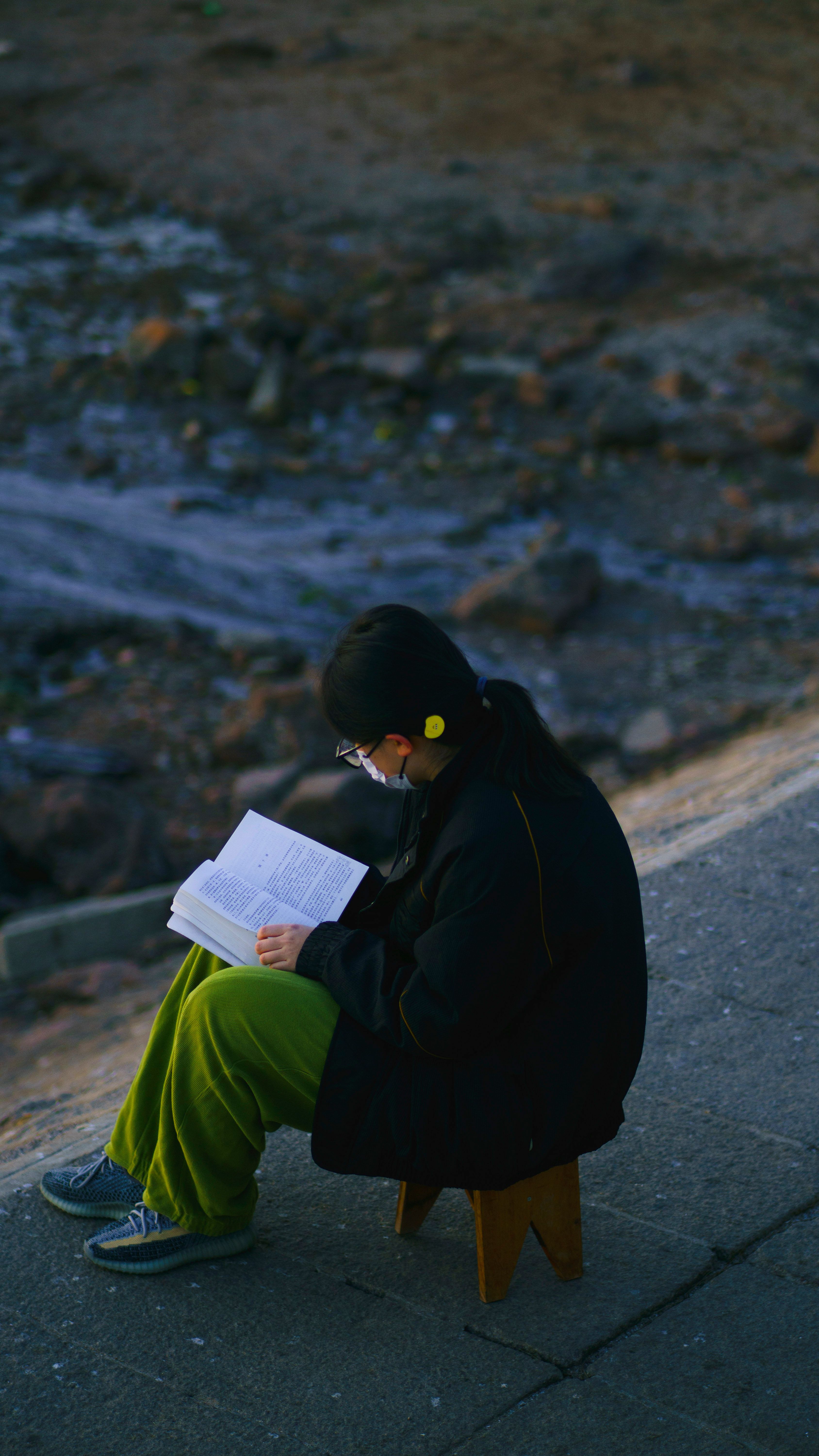A woman sits alone by the riverside in Qingdao, reading a book while wearing a face mask.