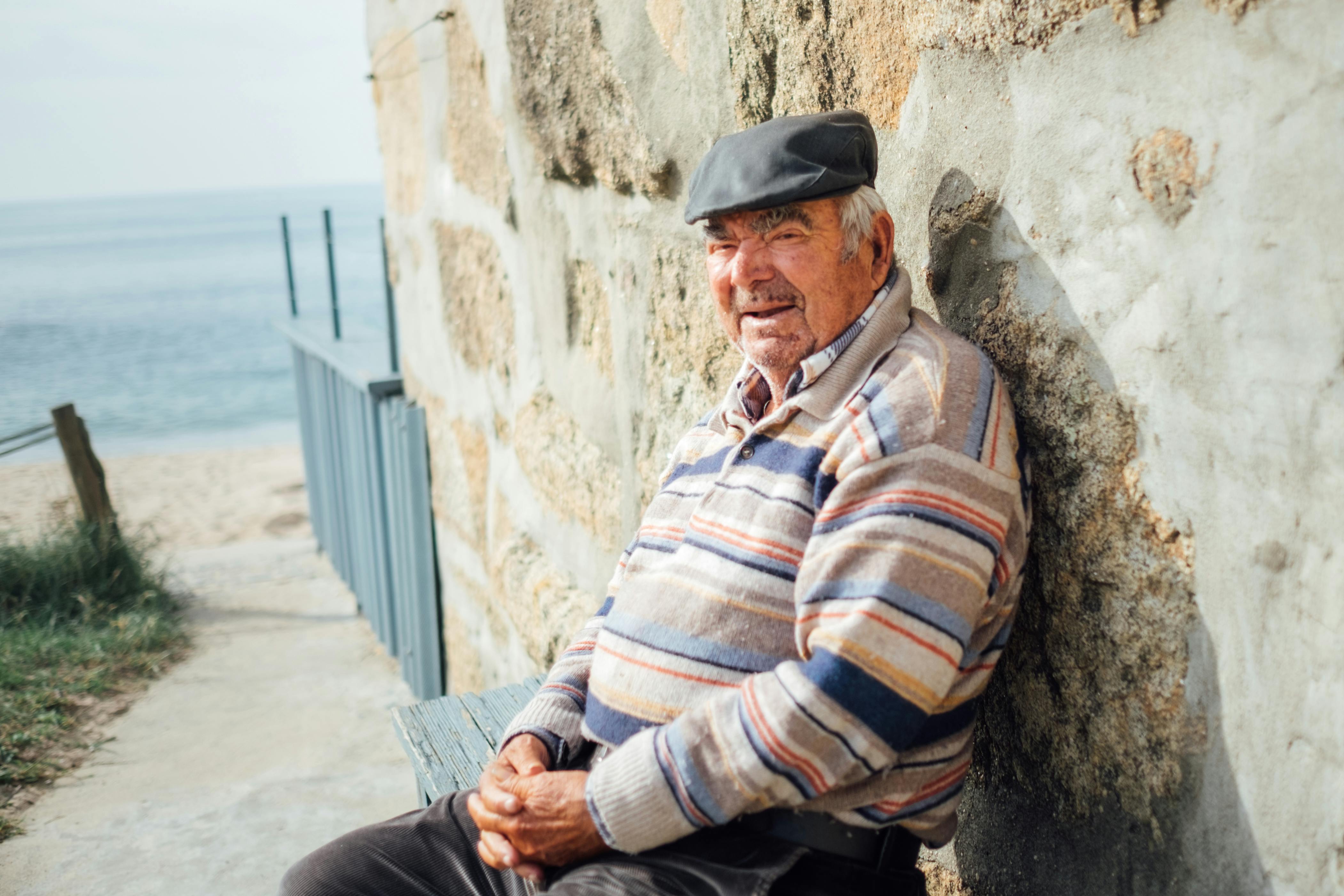 Senior man in striped sweater and cap sitting by a stone wall at the beach.