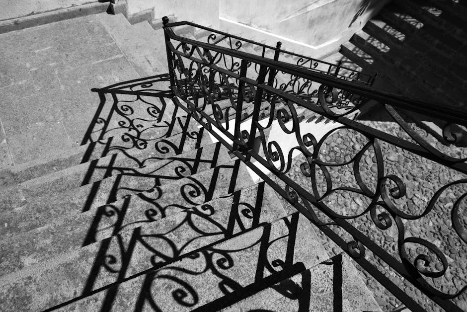 Captivating black and white image of wrought iron railings casting intricate shadows on stone steps in Ayvalık, Türkiye.