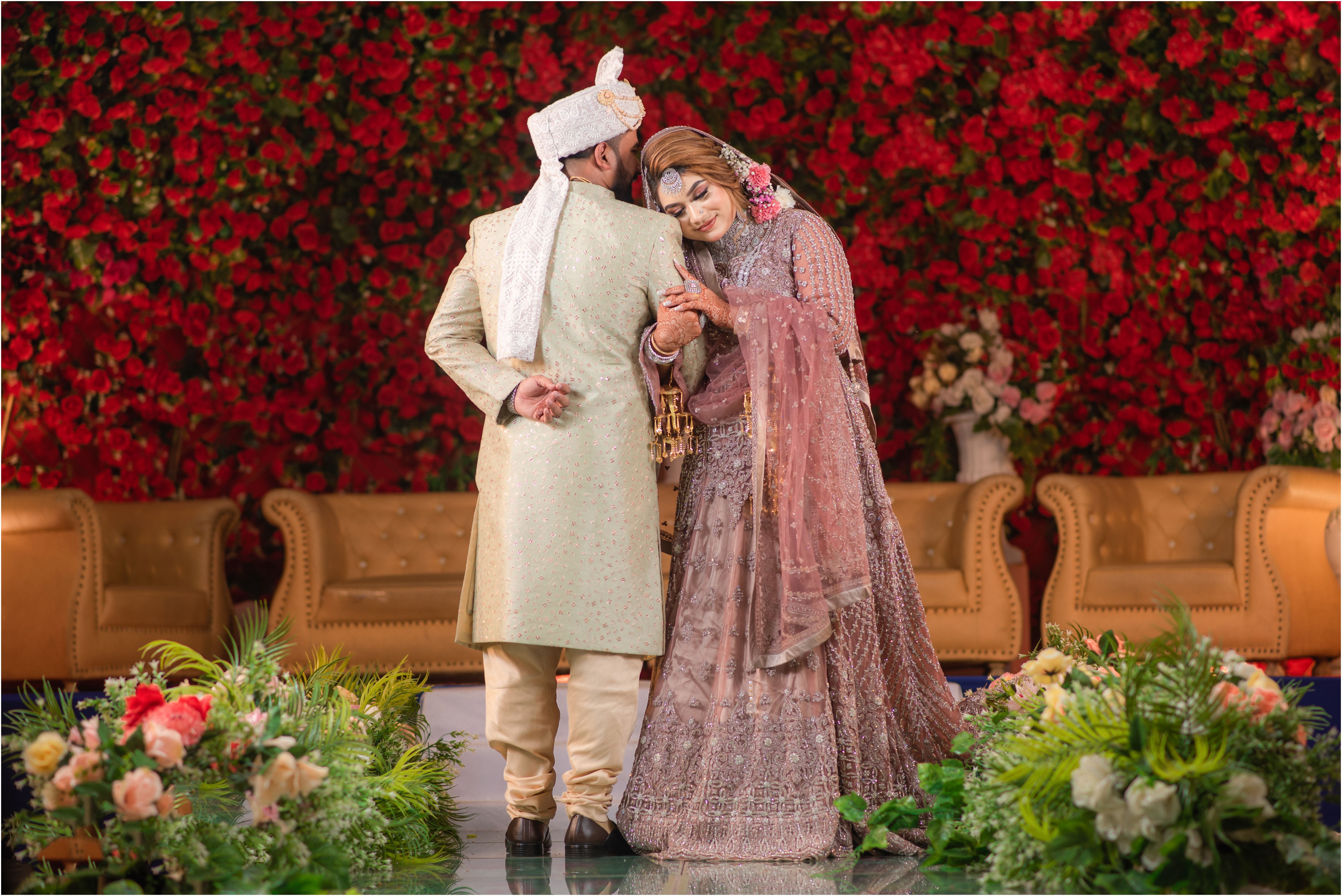 A joyful South Asian couple standing together in traditional wedding attire against a floral backdrop, celebrating their special day.