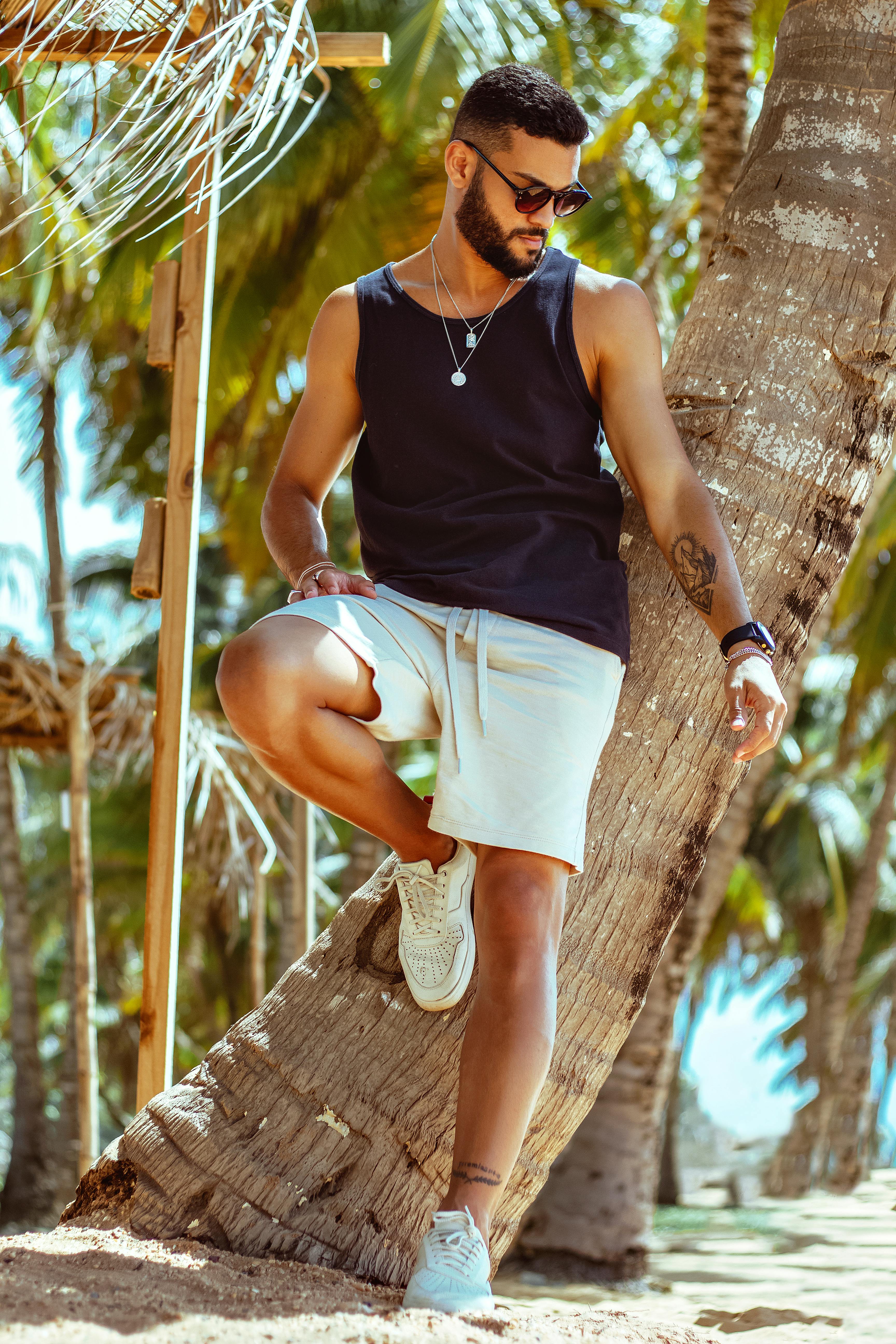 Man wearing sunglasses and casual attire posing by a palm tree on a sunny beach day.