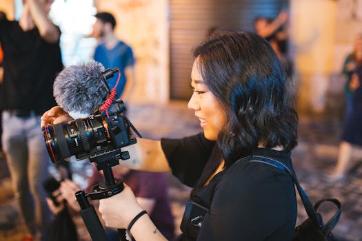 An Asian woman using a DSLR camera with a mic outdoors, engaged in filming or vlogging on a busy street.