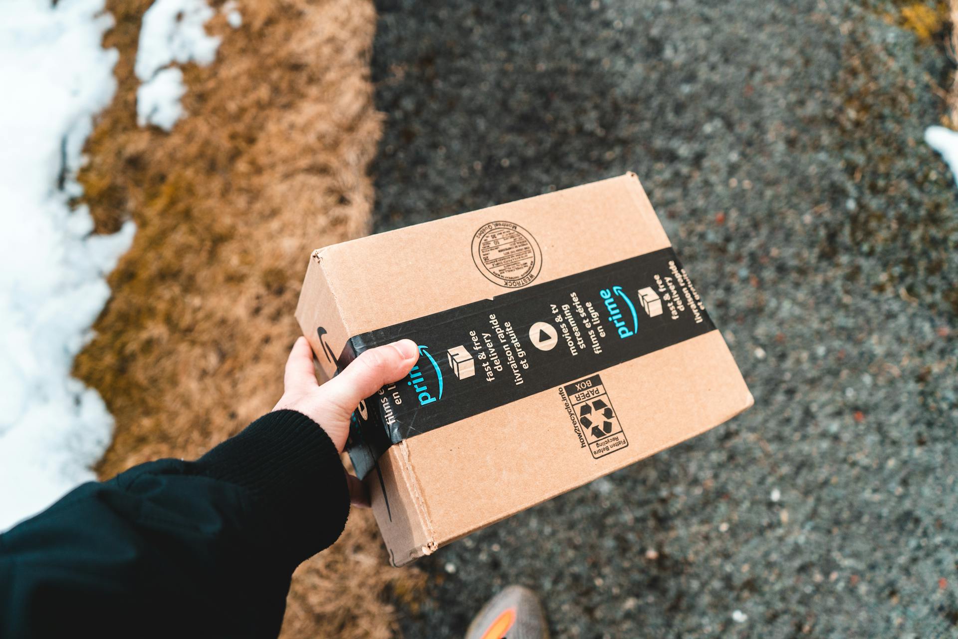 A person holding a cardboard Amazon Prime package on a snowy urban sidewalk.