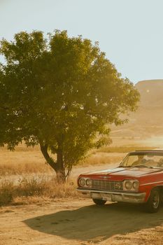 A vintage red car parked on a dusty road beside a lone tree in a warm afternoon landscape.