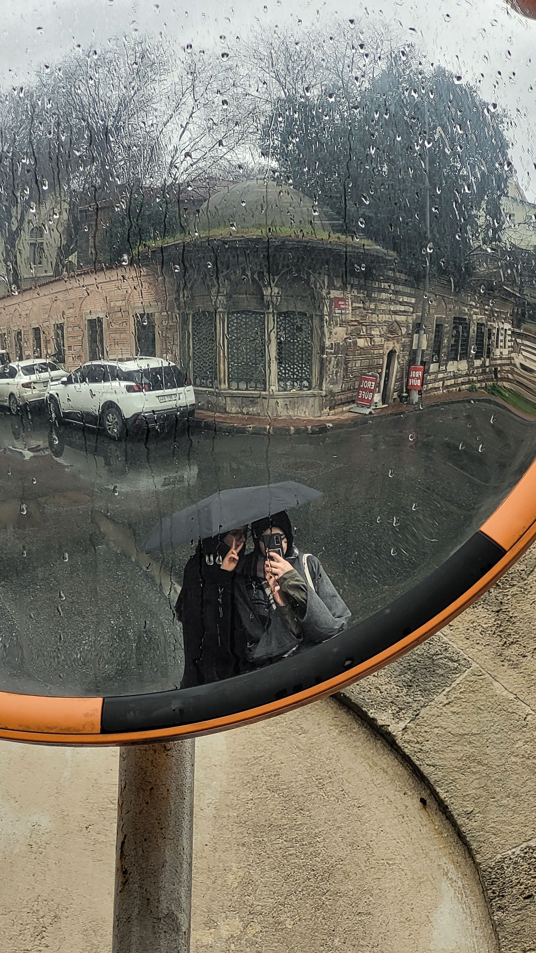 A person with an umbrella stands on a rainy day, reflected in a street mirror.