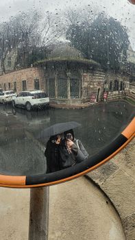A person with an umbrella stands on a rainy day, reflected in a street mirror.