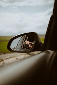 A creative reflection in a car mirror shows a person taking a photograph of scenic landscape.
