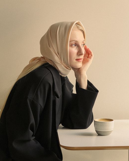 Young woman in hijab sitting indoors with a cup of coffee, looking thoughtful.