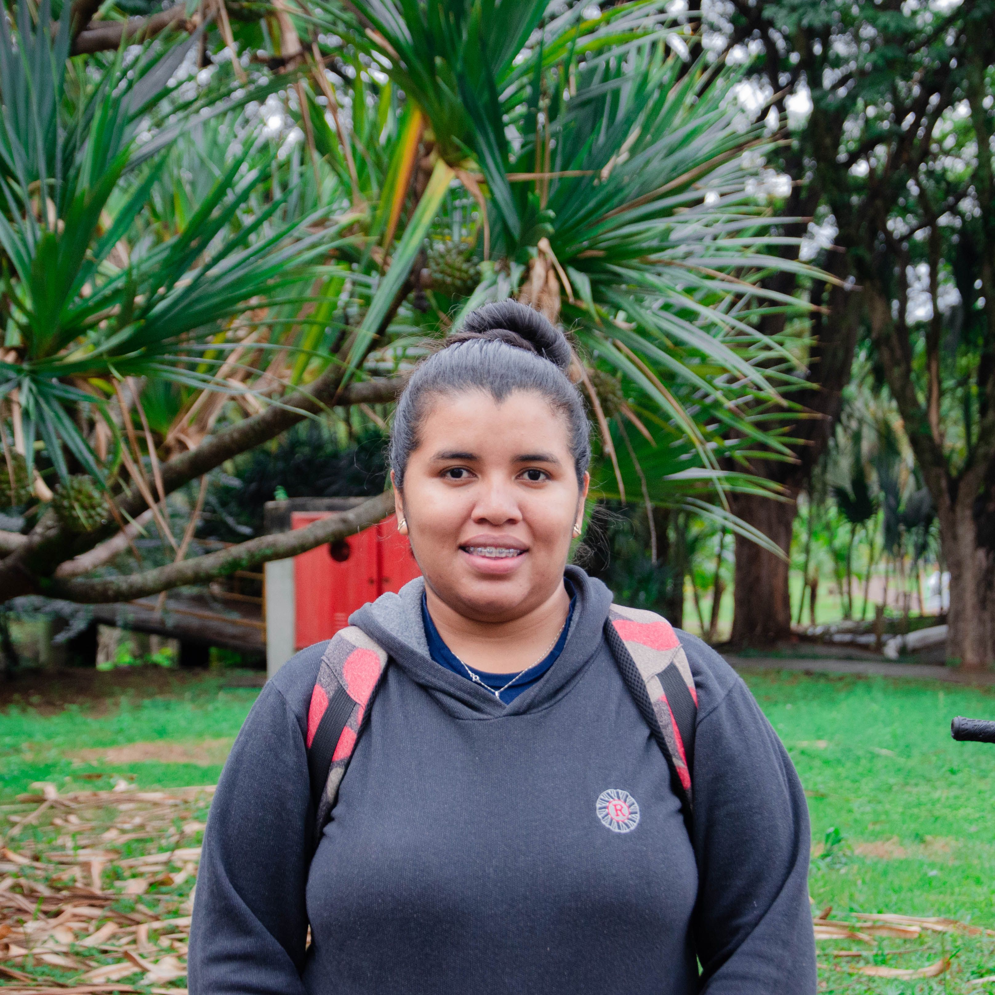 A woman in a hoodie with a backpack smiling in a verdant park with trees.