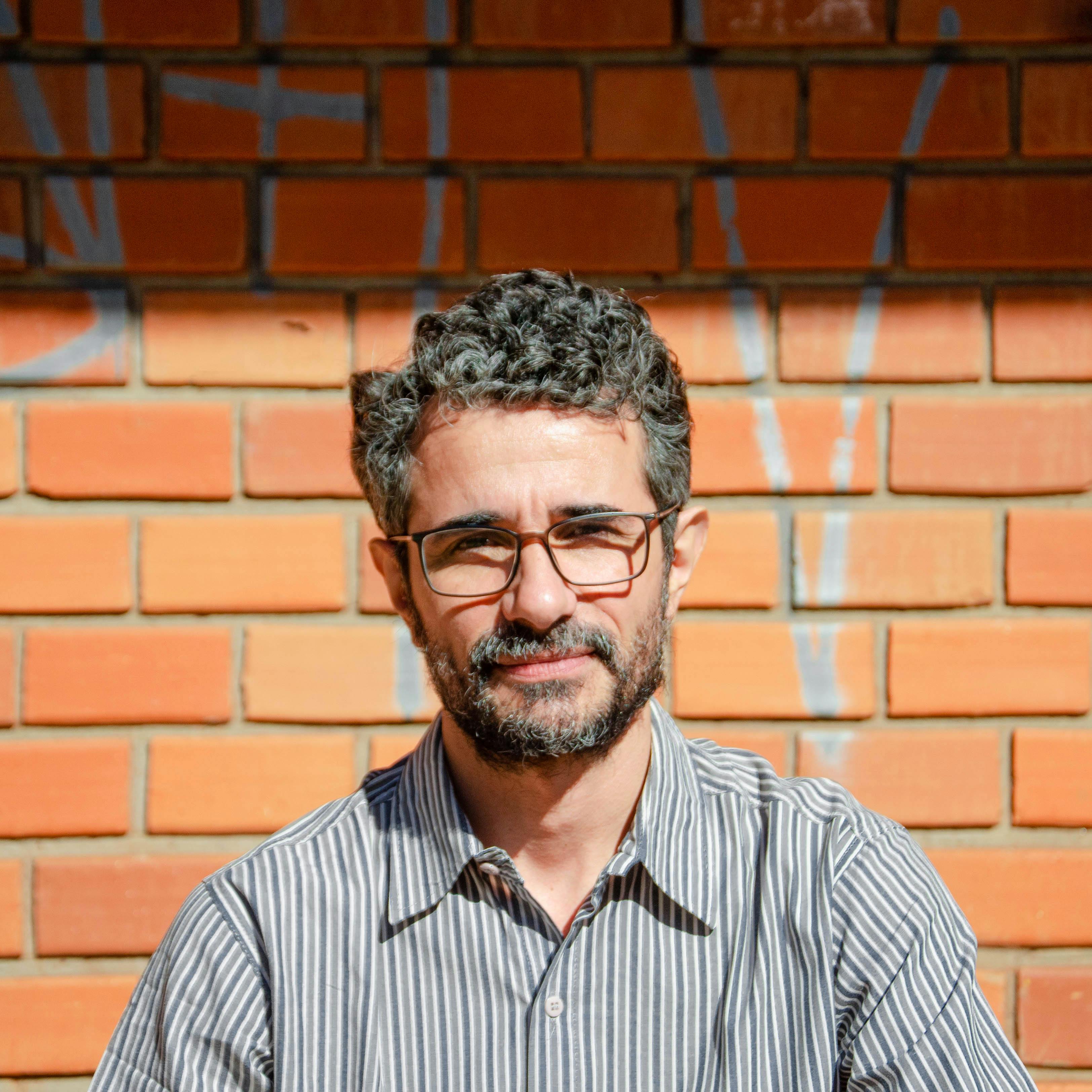 Outdoor portrait of a man with eyeglasses posing against a sunlit brick wall.