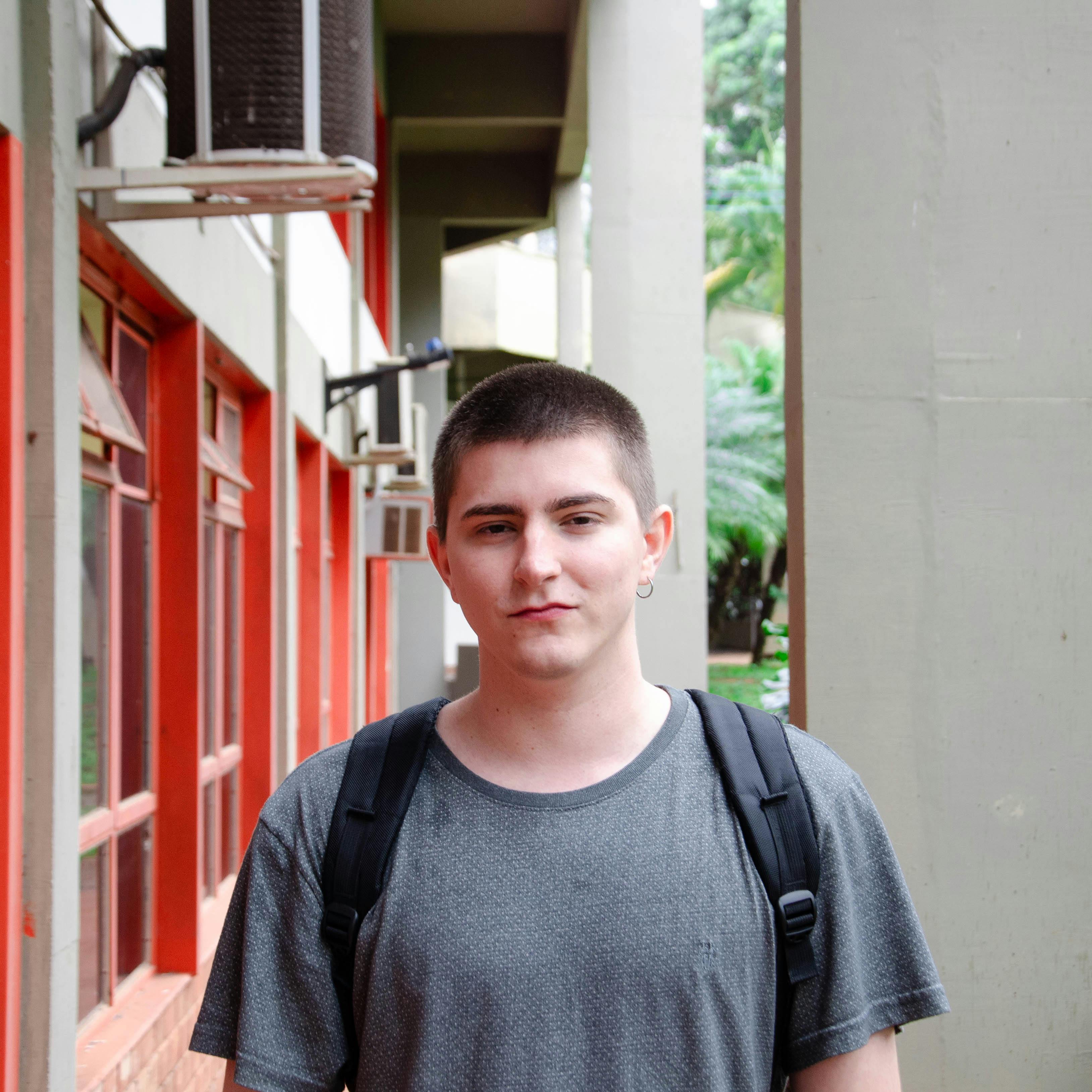 Portrait of a young man in a gray T-shirt standing outdoors against a building backdrop.