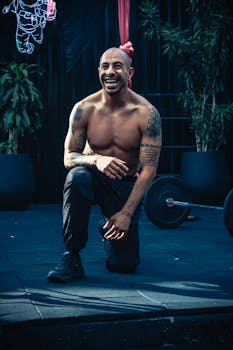 Smiling shirtless man kneeling while working out in an outdoor gym, exuding fitness and positivity.