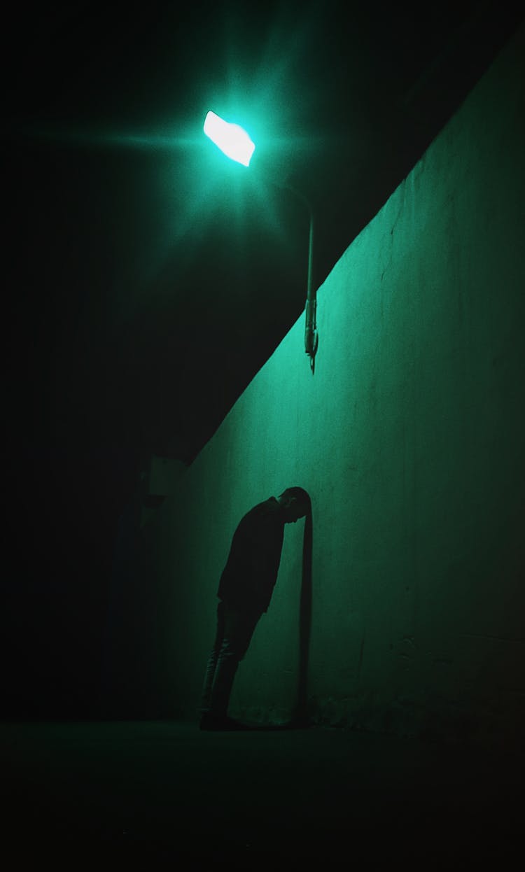 Silhouette Of A Man Leaning And Resting His Head Against The Wall Under A Lantern At Night 