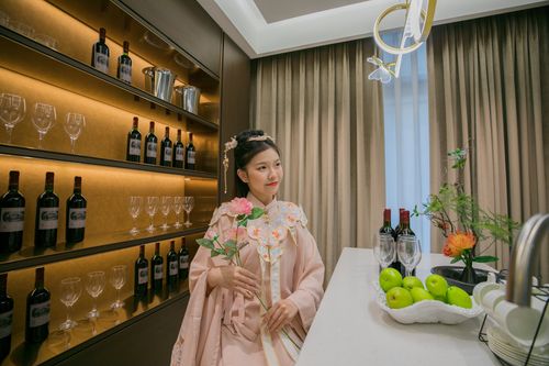Free A woman in traditional attire stands elegantly with a flower, surrounded by wine bottles and green apples indoors. Stock Photo