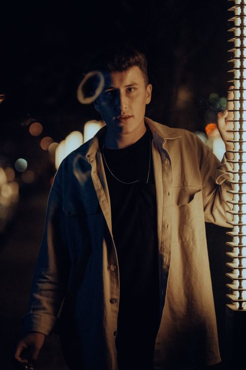 Free A man stands in an urban setting at night, illuminated by warm lights, wearing a trench coat. Stock Photo