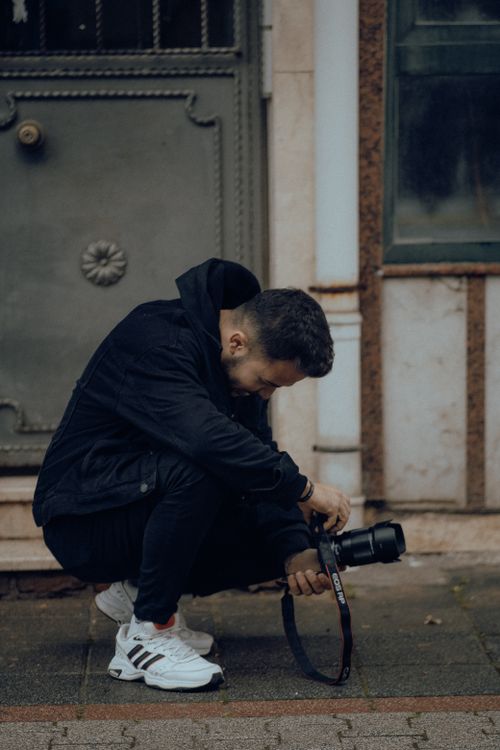 Free A man in black clothing squats on a city street, focusing on his camera. Stock Photo