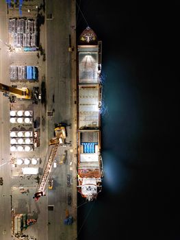 Top-down drone shot of an illuminated cargo ship docked at a bustling nighttime port.