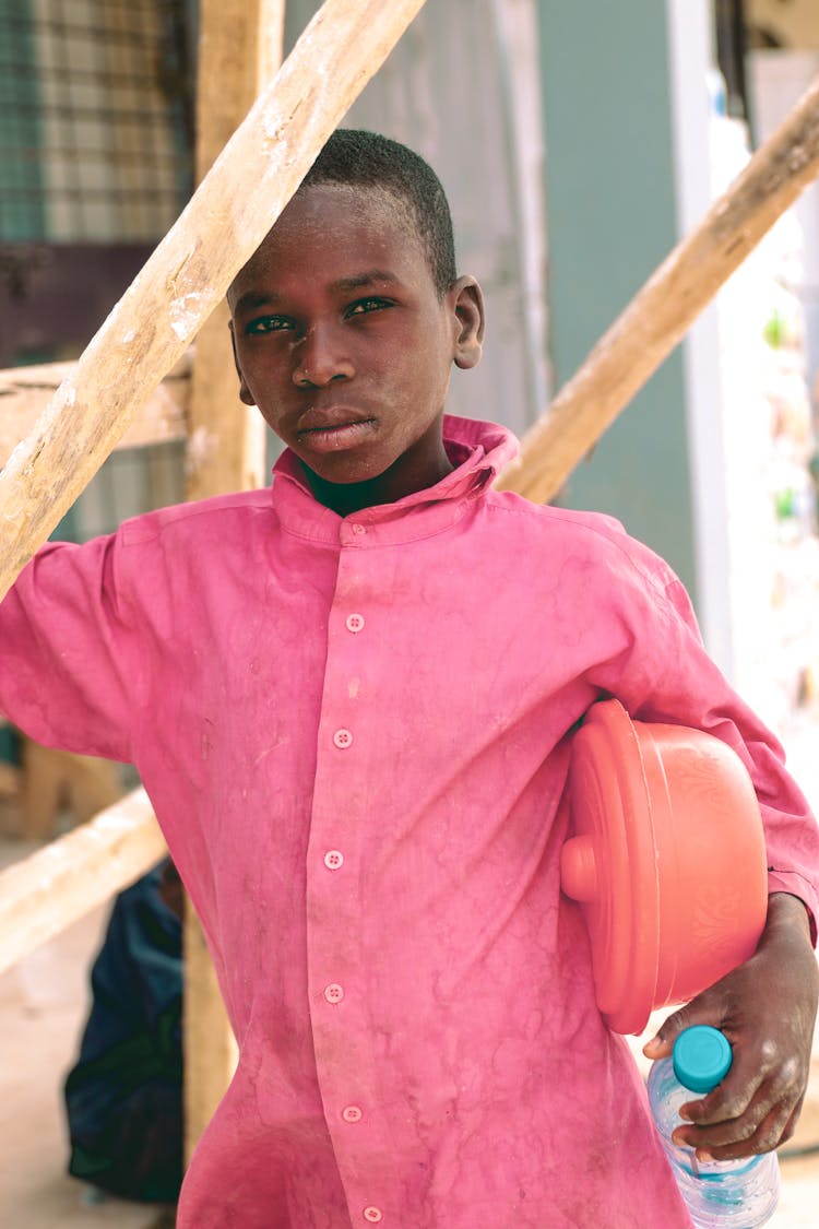 Boy In Pink Shirt Holding Pot Under Shoulder And Bottle Of Water In Hand