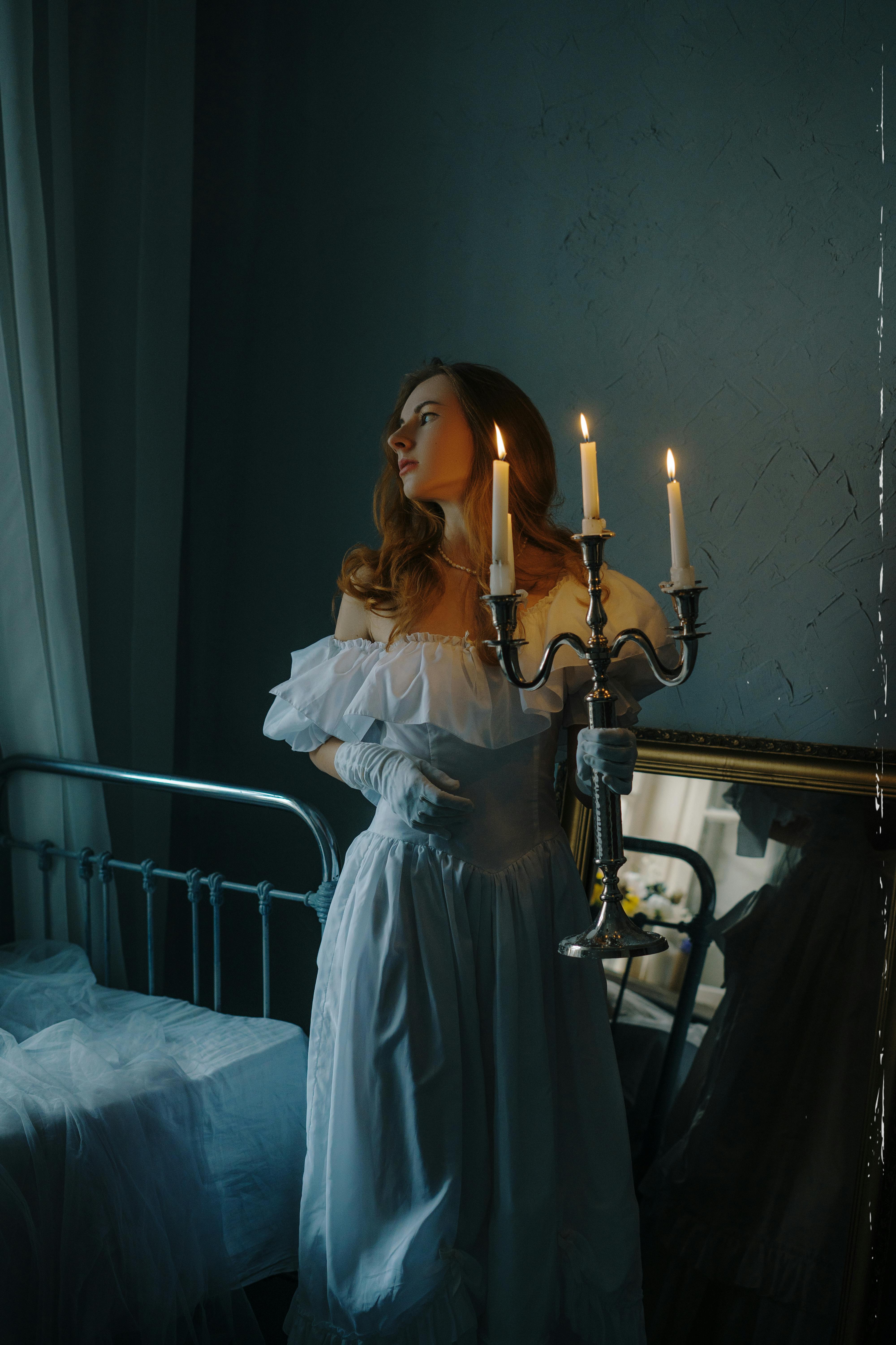 A woman in a white off-shoulder dress stands by a bed, holding a lit candlestick in a vintage room.