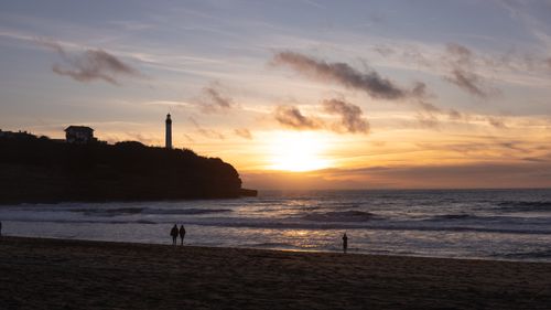 Free A peaceful sunset scene with silhouettes of people and a lighthouse on a beach. Stock Photo