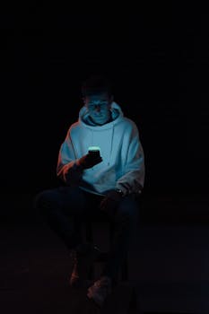Young man in a hoodie illuminated by smartphone light against dark background.