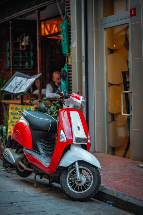 Free Vibrant red scooter parked on a bustling city street outside a cafe. Stock Photo