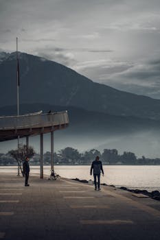 Two people walk along a lakeside promenade with mountains and cloudy skies.