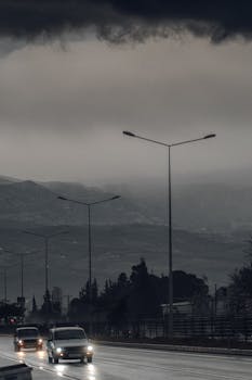 Cars driving on a wet road under dark overcast skies and streetlights, creating a dramatic scene.