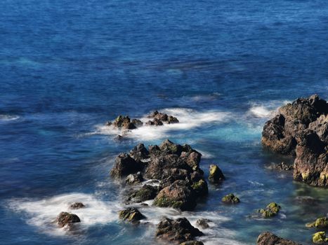 Dramatic view of sunlit eroded rocks along the coast, surrounded by a deep blue sea.