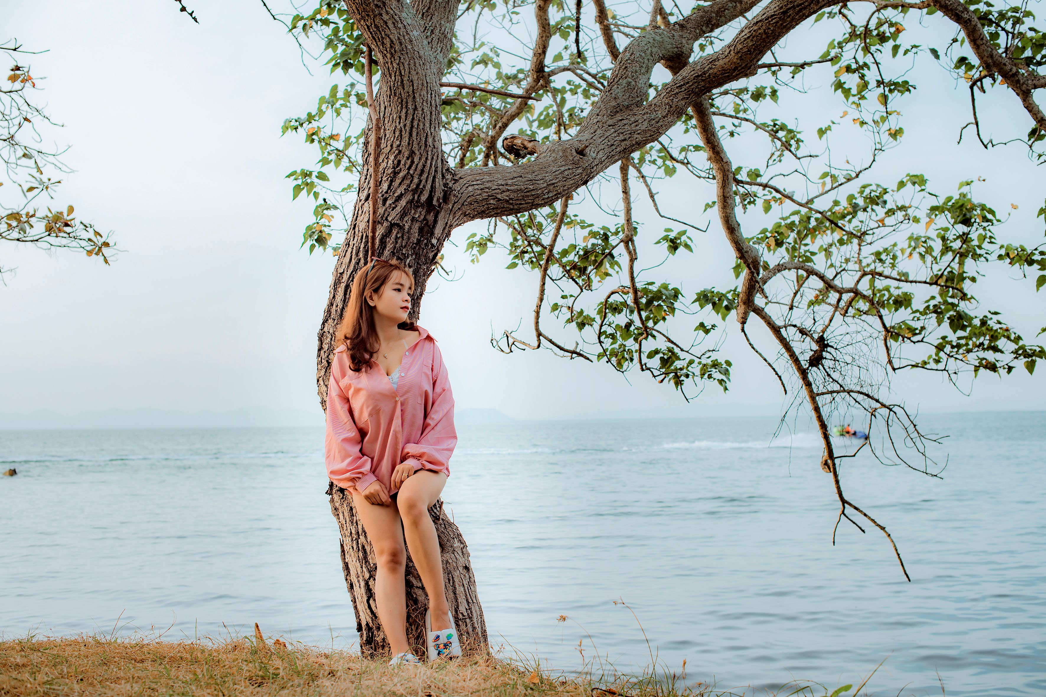 A woman in a pink sweater leans against a tree by the sea, enjoying a serene summer day.