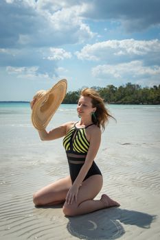 Woman in a swimsuit holding a straw hat on a sunny tropical beach.