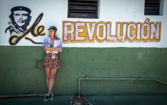 A woman in casual summer attire stands by a mural featuring Che Guevara and the word 'Revolución'.