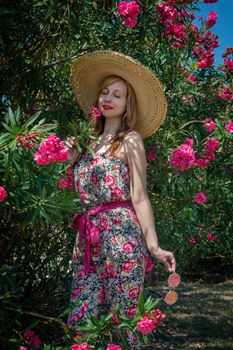 Woman in floral dress and straw hat, surrounded by blooming pink flowers on a sunny day.