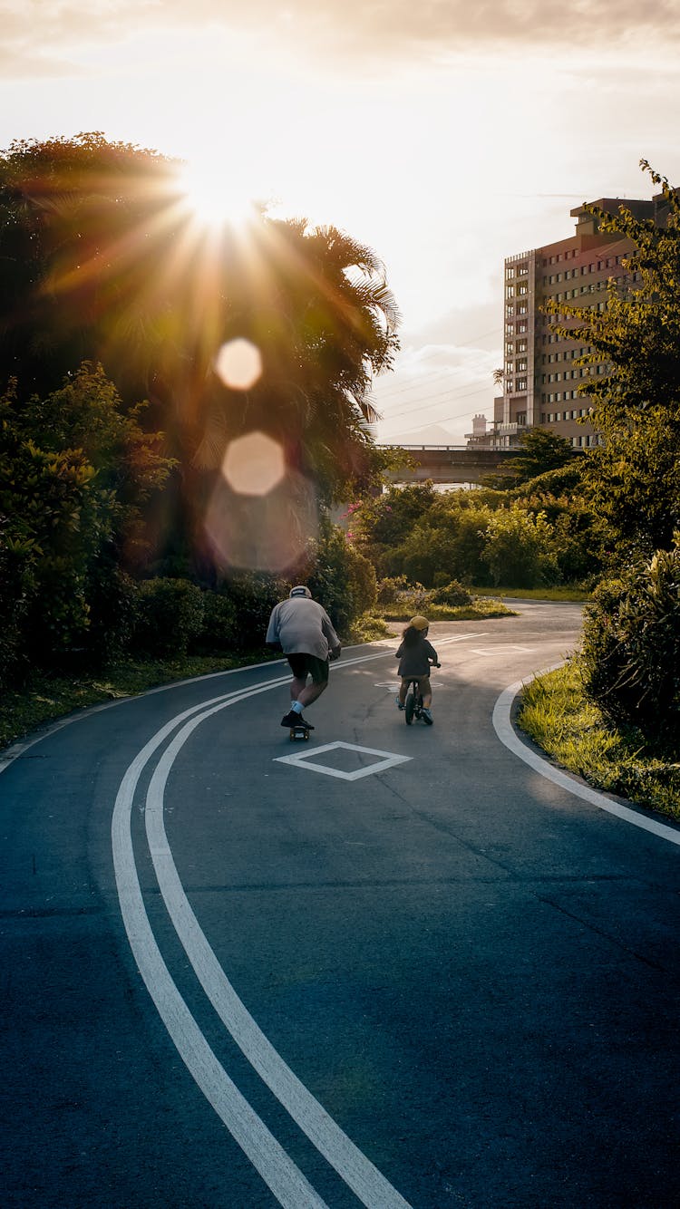 Father Skateboarding And Daughter Cycling At Park At Sunset