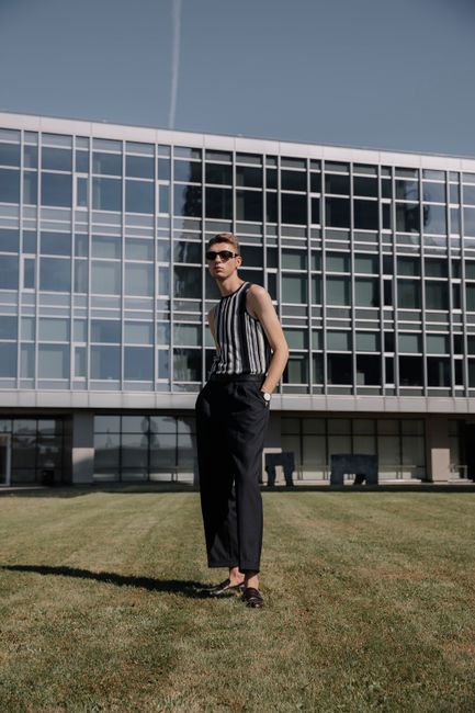 Man in striped top by a modern glass building, hands in pockets, sunny day.