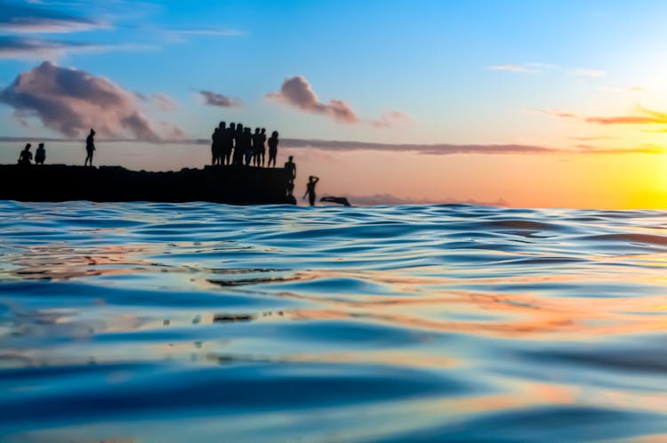 Close Up Of Water On Shore With People Standing Behind At Sunset