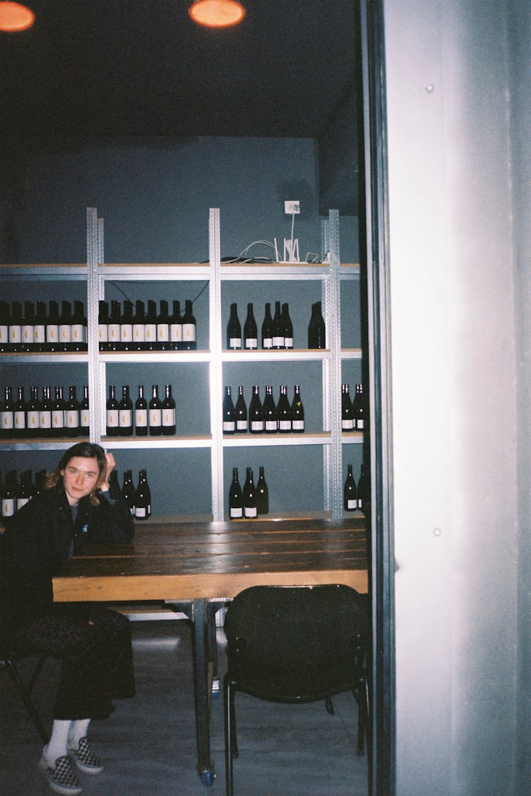 Woman Sitting By Table With Alcohol Bottles On Shelves Behind