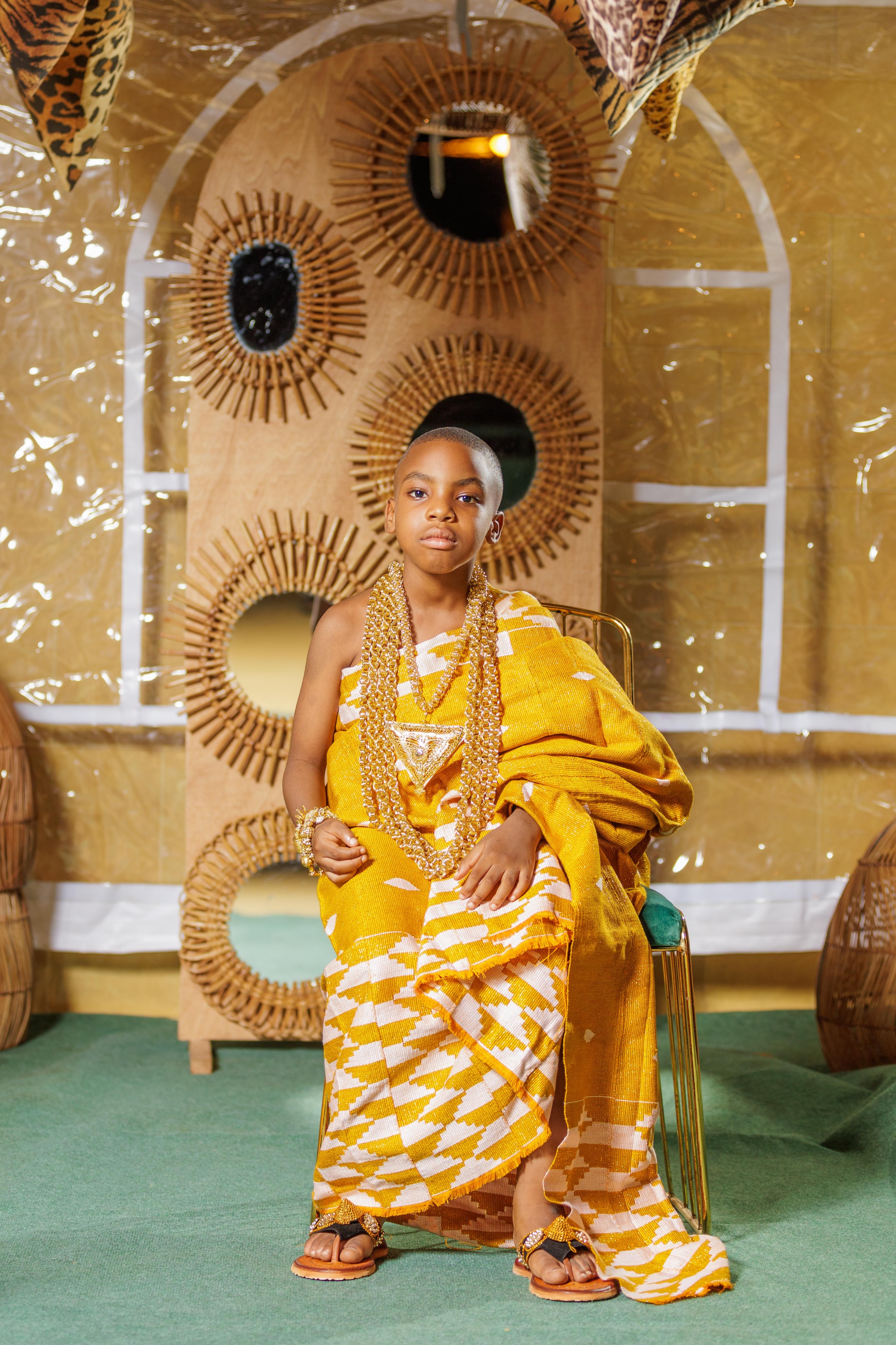 A young boy dressed in traditional clothing, sitting indoors with cultural decor.