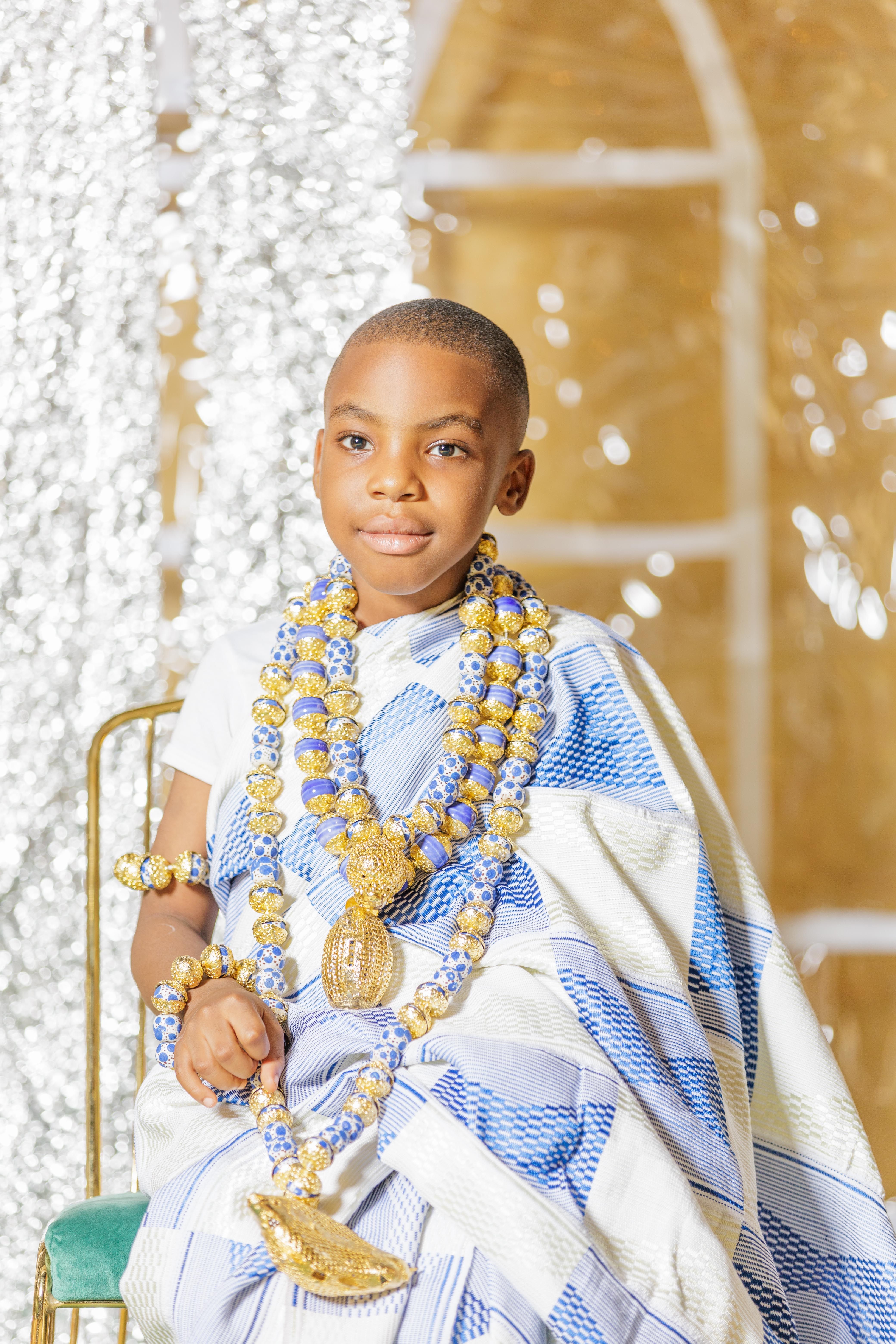 A young boy smiling, wearing traditional African clothing and jewelry, captured indoors.