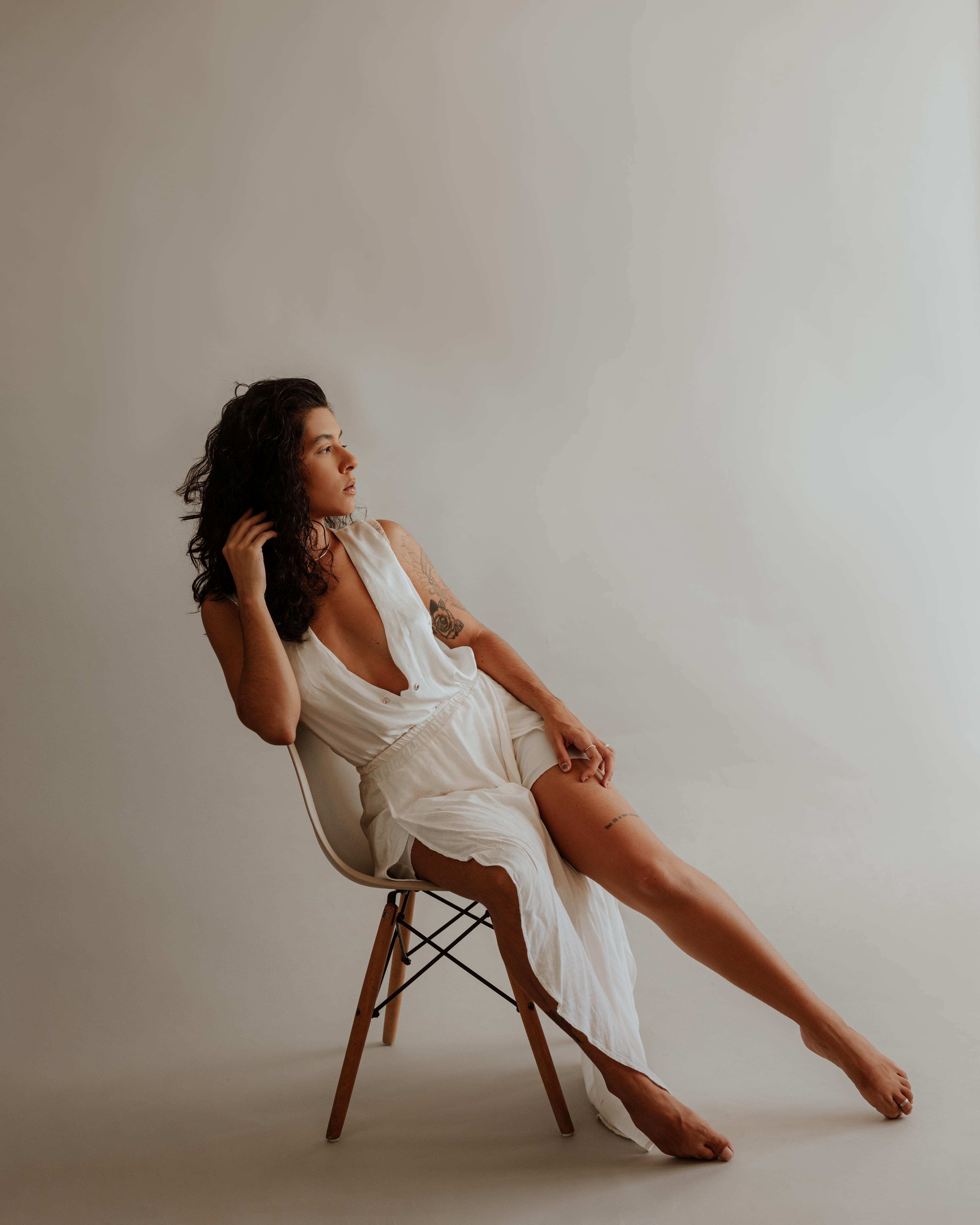 A stylish woman in a white dress sits gracefully on a chair in a studio setting.