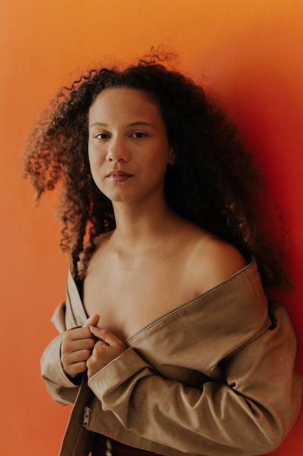 A portrait of a woman with curly hair and brown jacket against an orange background.