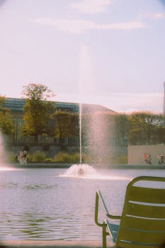 A peaceful fountain in a Paris park with people strolling and empty chairs nearby.