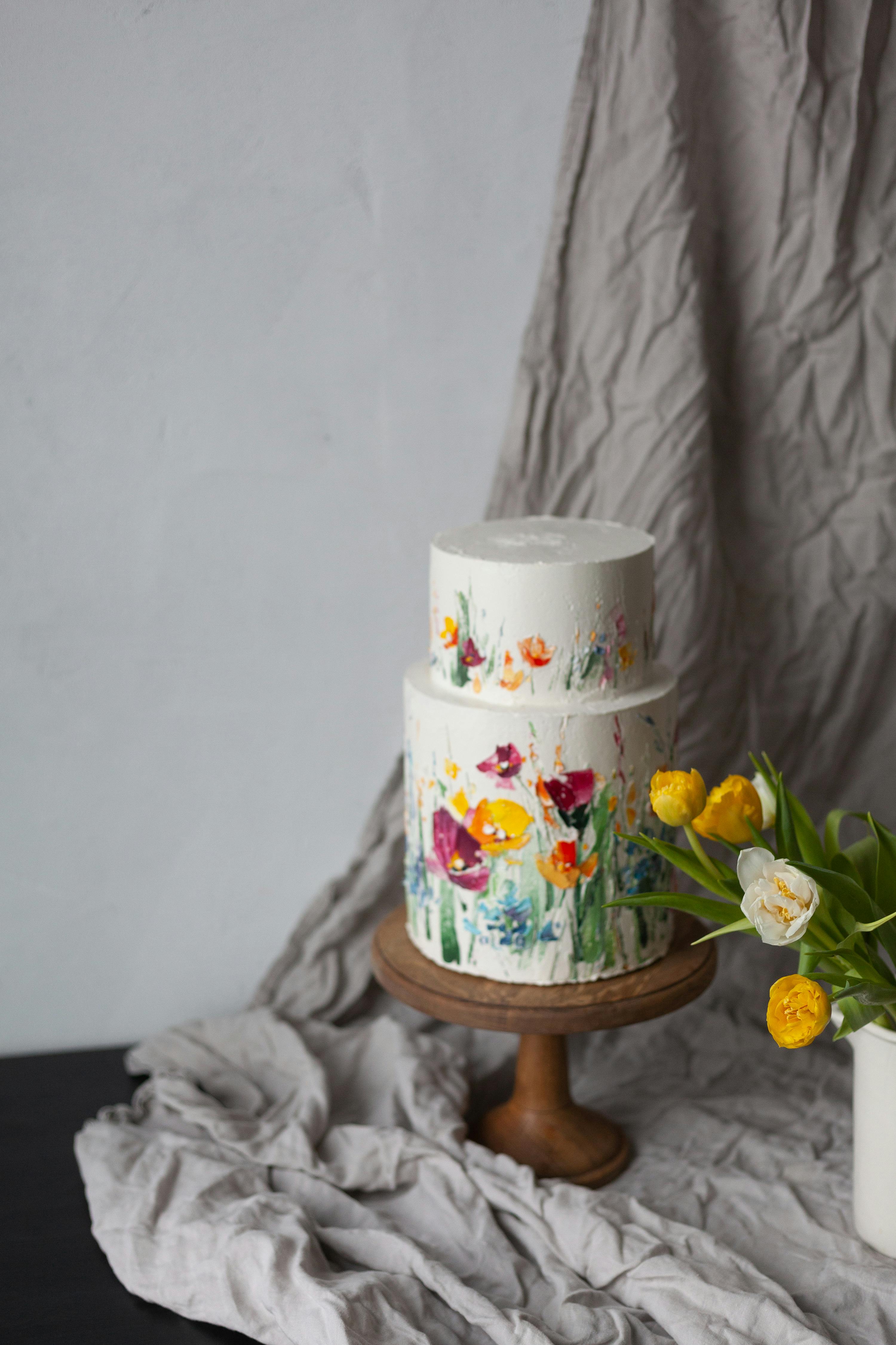 Elegant floral cake on wood stand with yellow tulips in vase.