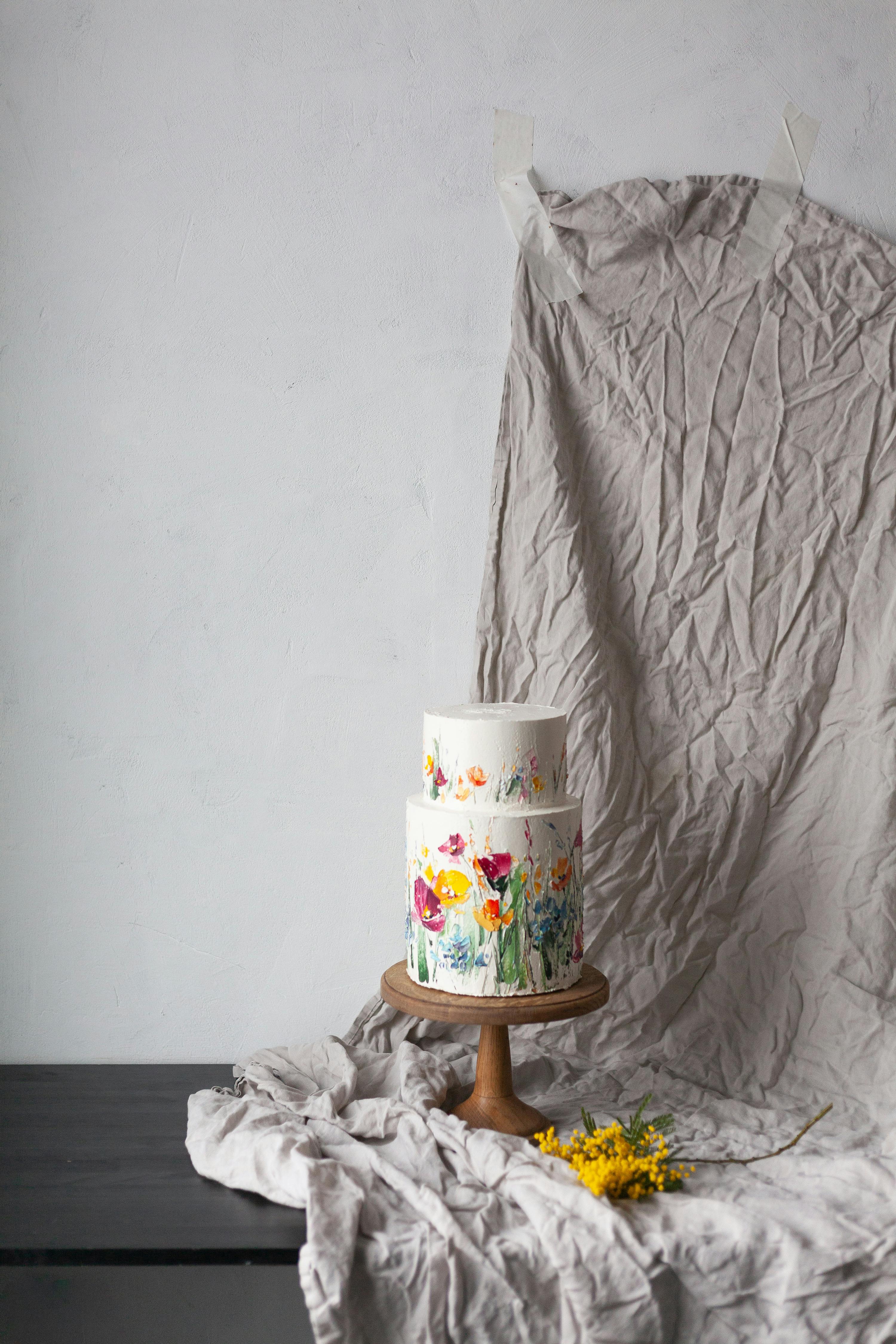 A two-tier floral cake elegantly displayed on a wooden cake stand with draped fabric backdrop.