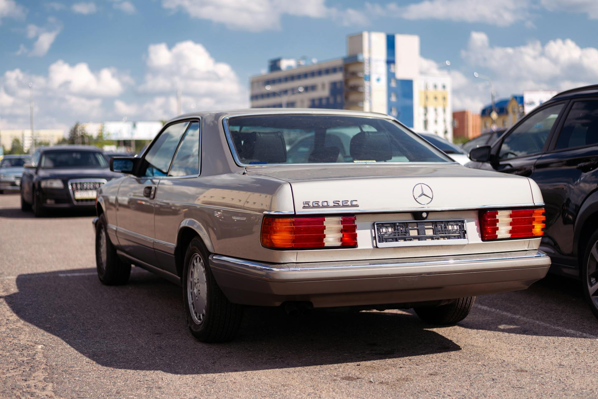 Classic Mercedes 560 SEC in a parking lot under a bright blue sky, showcasing vintage elegance.