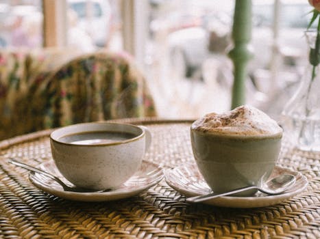 Two coffee cups with frothy cappuccino and black coffee on a wicker table in a cozy café setting.