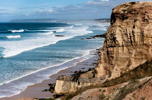 Free Stunning view of a rugged cliff overlooking a serene ocean with waves crashing on the sandy beach. Stock Photo