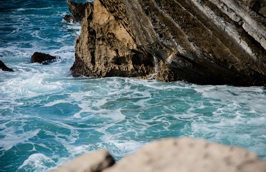 Waves crash against a dramatic rock formation on a vibrant blue ocean coast.