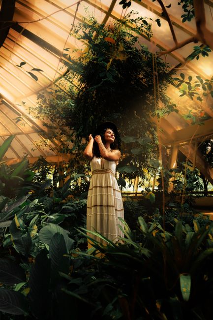 A young woman in a maxi dress embraces nature in a lush greenhouse setting in Belo Horizonte, Brazil.