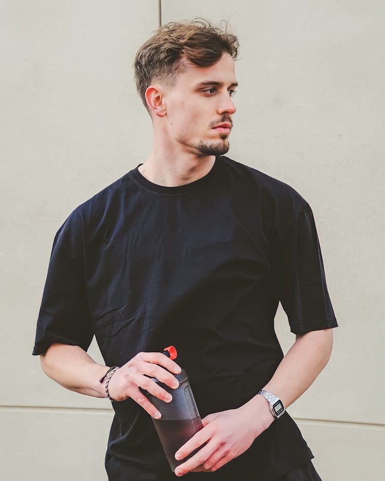 Young Man In A Black T-shirt Standing Outside And Holding A Bottle With A Drink 